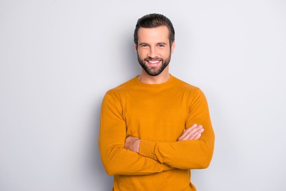 Ein Mann mit Stil im orangen Pullover lächelt und posiert mit verschränkten Armen vor grauem Hintergrund.