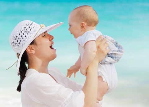 Stilsicher in wei&szlig; mit passendem Sommerhut gekleidet hebt eine Mutter ihr Baby in die Luft.