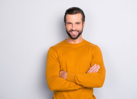 Ein Mann mit Stil im orangen Pullover l&auml;chelt und posiert mit verschr&auml;nkten Armen vor grauem Hintergrund.