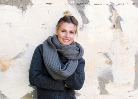 Eine Dame posiert in grauer Winterjacke mit passendem Schal l&auml;chelnd vor einer hellen Wand.